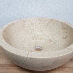 Pu&ograve; includere: Un lavabo da bagno rotondo in marmo bianco con un foro di scarico al centro. Il lavabo ha una superficie liscia e lucida con venature visibili.