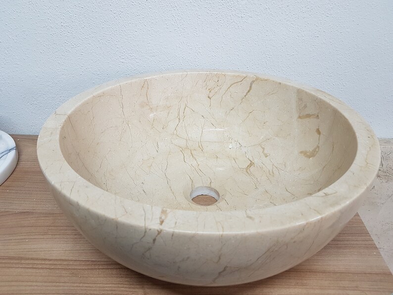 Pu&ograve; includere: Un lavabo da bagno rotondo in marmo bianco con un foro di scarico al centro. Il lavabo ha una superficie liscia e lucida con venature visibili.