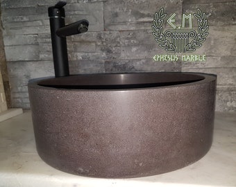 May include: A round, dark brown stone vessel sink with a black faucet. The sink is made of natural stone and has a rough, textured surface. The faucet is tall and slender, with a single handle. The sink is set against a backdrop of gray stone tiles.
