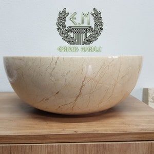 Pu&ograve; includere: Un grande lavabo rotondo in marmo color crema con venature marroni. Il lavabo &egrave; posto su un piano di lavoro in legno. Il testo "E.M.  GRECUS MARBLE" &egrave; visibile nella parte superiore centrale dell'immagine.