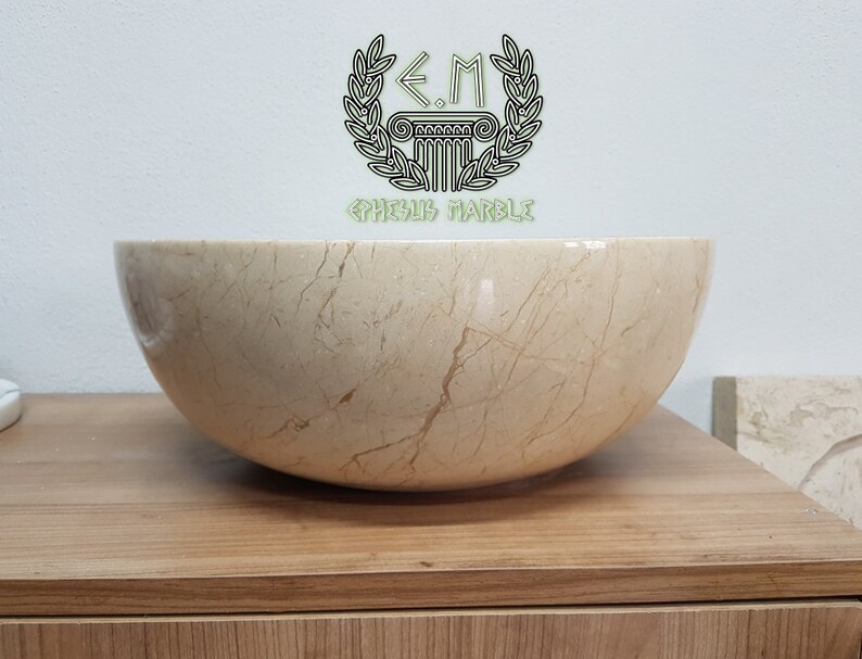 Pu&ograve; includere: Un grande lavabo rotondo in marmo color crema con venature marroni. Il lavabo &egrave; posto su un piano di lavoro in legno. Il testo "E.M.  GRECUS MARBLE" &egrave; visibile nella parte superiore centrale dell'immagine.