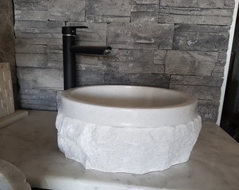 May include: White marble sink with a rough, textured exterior, resembling a rock formation. A black faucet is mounted on the wall behind the sink.