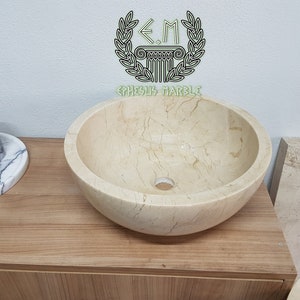 Pu&ograve; includere: Un lavabo rotondo in marmo color crema con una superficie liscia e lucida. Il lavabo presenta una sottile venatura e un piccolo foro di scarico al centro. Il lavabo &egrave; posizionato su un piano di lavoro in legno.