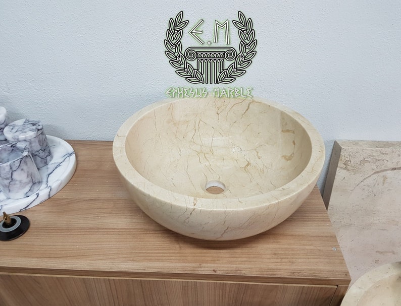 Pu&ograve; includere: Un lavabo rotondo in marmo color crema con una superficie liscia e lucida. Il lavabo presenta una sottile venatura e un piccolo foro di scarico al centro. Il lavabo &egrave; posizionato su un piano di lavoro in legno.