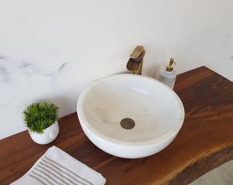 May include: A white marble vessel sink with a gold faucet and a white soap dispenser on a wooden countertop. A small potted plant and a white towel are also on the countertop.