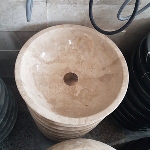 Pu&ograve; includere: Primo piano di un lavabo da bagno in marmo beige con un tappo di scarico in ottone. Il lavabo &egrave; impilato sopra altri lavabi.