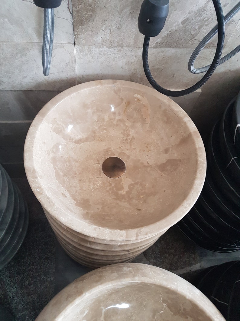 Pu&ograve; includere: Primo piano di un lavabo da bagno in marmo beige con un tappo di scarico in ottone. Il lavabo &egrave; impilato sopra altri lavabi.