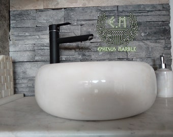May include: White marble vessel sink with a black faucet and a white marble soap dispenser. The sink is on a white marble countertop. The background is a gray stone wall with the text "E.M. Ephesus Marble".