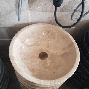 Pu&ograve; includere: Un lavabo rotondo in marmo beige con un tappo di scarico in ottone. Il lavabo &egrave; posto su un piedistallo ed &egrave; circondato da altri lavabi.