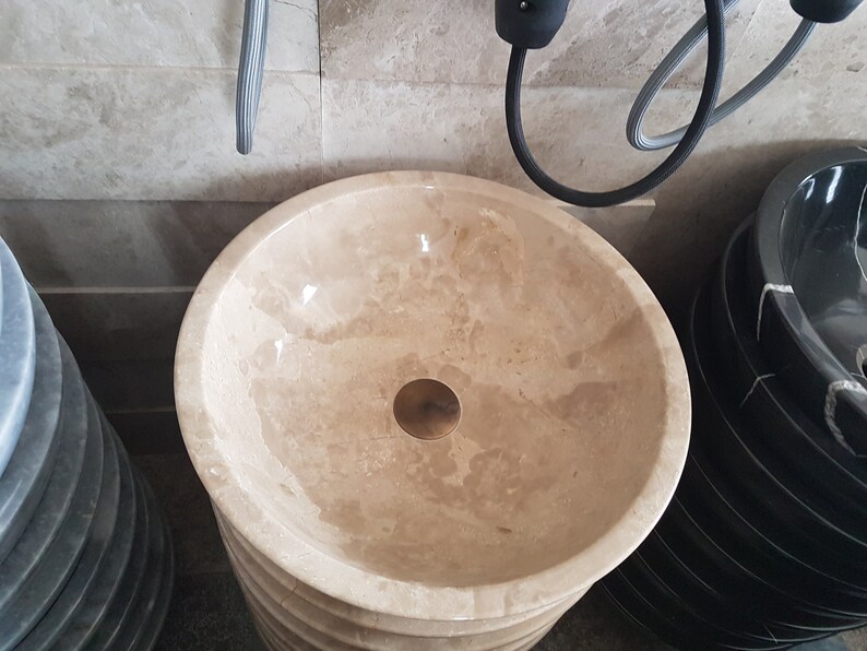 Pu&ograve; includere: Un lavabo rotondo in marmo beige con un tappo di scarico in ottone. Il lavabo &egrave; posto su un piedistallo ed &egrave; circondato da altri lavabi.
