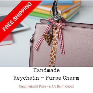 May include: A pink purse with a silver handle is adorned with a handmade keychain. The keychain features a pizza slice charm, a red and white gingham ribbon, and a pizza-patterned strap. The text "Handmade Keychain - Purse Charm" is visible.