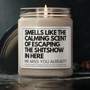 May include: A beige soy candle with a black label that reads "Smells like the calming scent of escaping the shitshow in here. We miss you already! Soy candle made in USA. You're looking good."