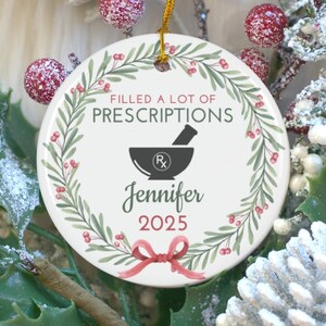 May include: A round, white ceramic ornament with the text "FILLED A LOT OF PRESCRIPTIONS" and a mortar and pestle graphic. The name "Jennifer" and the year "2025" are also displayed. The ornament is decorated with a green wreath and a red bow.