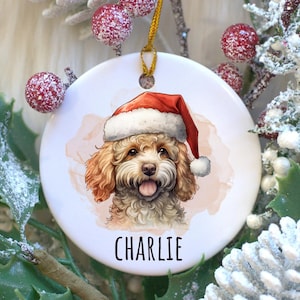 May include: A round Christmas ornament featuring a watercolor illustration of a dog wearing a Santa hat. The dog's name, "Charlie," is printed below the image. The ornament hangs from a gold string and is surrounded by festive greenery.