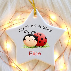 Puede incluir: Un adorno blanco en forma de estrella con una mariquita de dibujos animados y el texto "CUTE AS A BUG!" y el nombre "Elise". El adorno está decorado con una cuerda dorada y luces de hadas.