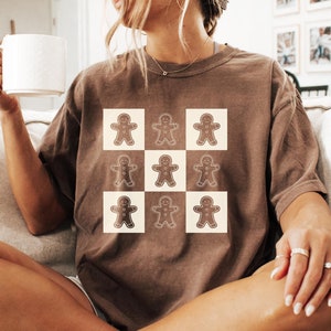 May include: A brown t-shirt featuring a grid of gingerbread man graphics. The gingerbread men are brown with white icing details, set against a cream-colored square background. The shirt is a relaxed fit, with short sleeves.