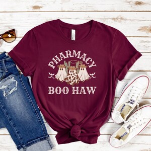 May include: A maroon t-shirt with a white graphic of three ghosts wearing cowboy hats and the text "Pharmacy Boo Haw".