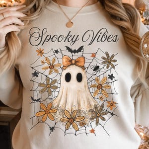 May include: Beige sweatshirt with a Halloween-themed graphic. The design features a ghost with a bow, surrounded by a spider web, flowers, and the words "Spooky Vibes". The sweatshirt is a neutral color.