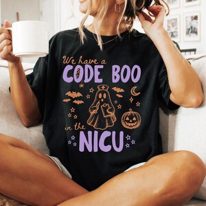 May include: A black t-shirt featuring a graphic with the text "We have a CODE BOO in the NICU" in purple and orange. The design includes a ghost wearing a nurse cap, a pumpkin, bats, stars, and a crescent moon in orange line art.