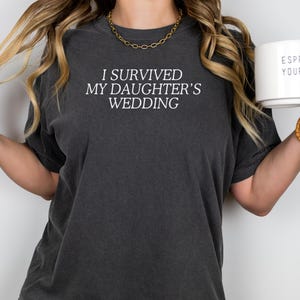 May include: A woman wearing a dark grey t-shirt with the text "I survived my daughter's wedding" in white. She is holding a white mug with the text "Espresso Yourself" in black.