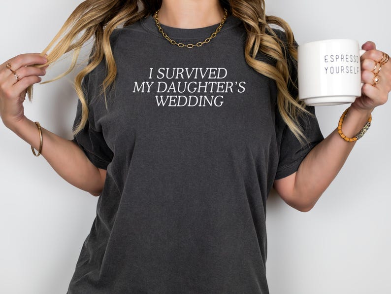 May include: A woman wearing a dark grey t-shirt with the text "I survived my daughter's wedding" in white. She is holding a white mug with the text "Espresso Yourself" in black.
