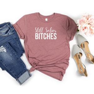 May include: A mauve t-shirt with the text "Still Sober Bitches" printed in white.