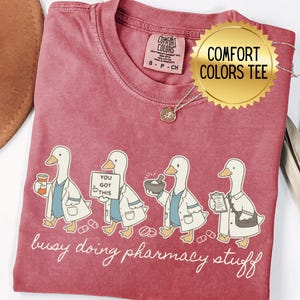 May include: A red Comfort Colors tee shirt featuring a graphic of four cartoon ducks in lab coats, each carrying pharmacy items. The text "busy doing pharmacy stuff" is below the ducks. A gold pendant necklace and sticker are visible.