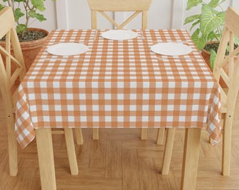 May include: A square orange and white checkered tablecloth covers a wooden table set for a meal. Three white plates are on the table. Two wooden chairs are on either side of the table. Potted plants are in the background.