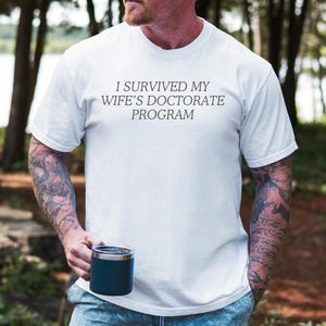 May include: A white t-shirt with the text "I survived my wife's doctorate program" printed on the front. The person wearing the shirt is standing in a forest.
