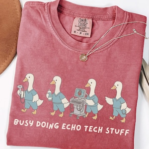 May include: Red Comfort Colors t-shirt featuring four cartoon ducks dressed as medical professionals. The ducks are depicted with medical equipment and the text "BUSY DOING ECHO TECH STUFF" is printed below. A gold necklace is draped across the shirt.