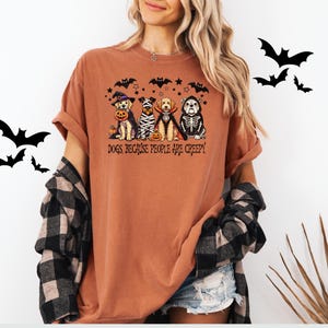 May include: Terracotta-colored t-shirt featuring a Halloween-themed graphic. The graphic depicts four dogs dressed in costumes, including a witch, mummy, vampire, and skeleton. The text "Dogs Because People Are Creepy" is printed below the dogs.