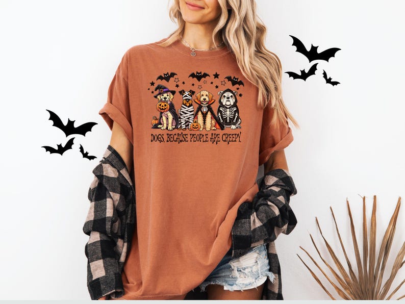 May include: Terracotta-colored t-shirt featuring a Halloween-themed graphic. The graphic depicts four dogs dressed in costumes, including a witch, mummy, vampire, and skeleton. The text "Dogs Because People Are Creepy" is printed below the dogs.