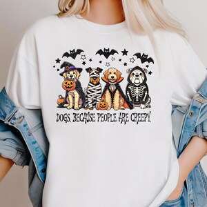 May include: White t-shirt featuring a Halloween-themed graphic. The design includes four dogs dressed in costumes, bats, stars, and the text "DOGS BECAUSE PEOPLE ARE CREEPY."