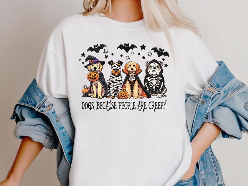 May include: White t-shirt featuring a Halloween-themed graphic. The design includes four dogs dressed in costumes, bats, stars, and the text "DOGS BECAUSE PEOPLE ARE CREEPY."