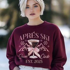 Puede incluir: Una sudadera color granate con un diseño gráfico rosa de una copa de cóctel con un lazo rosa y el texto "Après Ski Social Club Est. 2025".