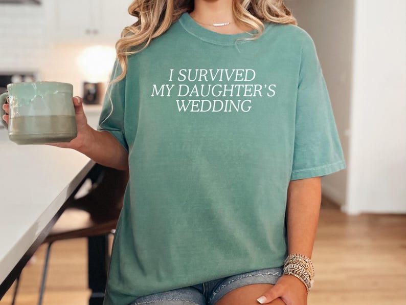 May include: A teal blue t-shirt with the text "I survived my daughter's wedding" in white.
