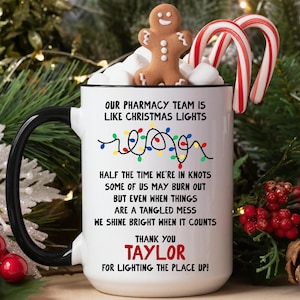May include: A white ceramic mug with a black handle and rim, featuring a graphic of colorful Christmas lights and text that reads: Our pharmacy team is like Christmas lights, half the time we're in knots, some of us may burn out, but even when things are a tangled mess, we shine bright when it counts, thank you Taylor for lighting the place up.