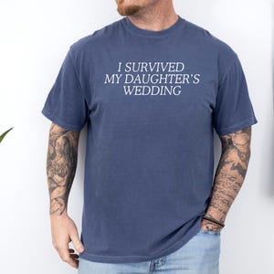 May include: A navy blue t-shirt with the text "I survived my daughter's wedding" printed in white.