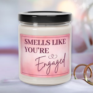 May include: A white candle in a glass jar with a black lid. The label on the jar is pink with white text that reads "Smells Like You're Engaged" with two small hearts above the word "Engaged".