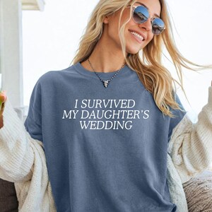 May include: A woman wearing a blue t-shirt with the text "I survived my daughter's wedding" in white. She is smiling and holding a glass of lemonade.