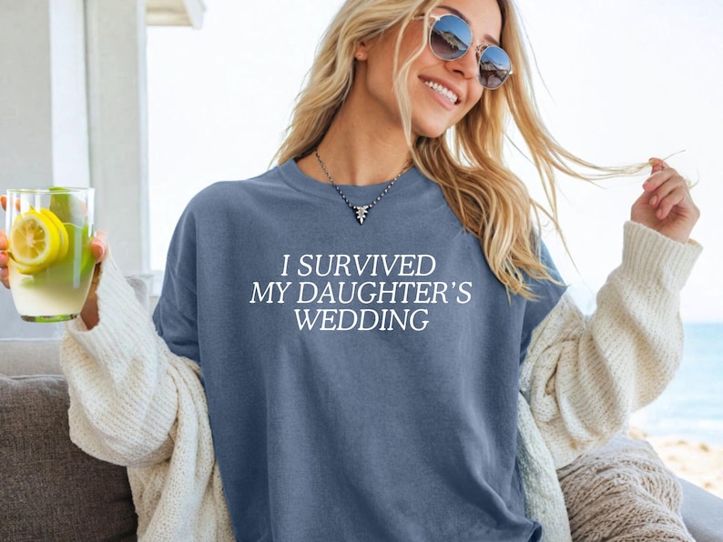 May include: A woman wearing a blue t-shirt with the text "I survived my daughter's wedding" in white. She is smiling and holding a glass of lemonade.