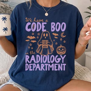 May include: A navy blue t-shirt featuring a graphic with the text We have a CODE BOO in the RADIOLOGY DEPARTMENT in purple and orange. The design includes a ghost holding an X-ray, bats, stars, a crescent moon, and a jack-o'-lantern pumpkin.