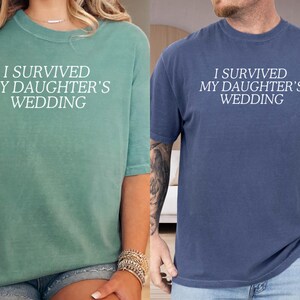 May include: Two t-shirts with the text "I survived my daughter's wedding" in white lettering. The shirt on the left is teal green and the shirt on the right is navy blue.