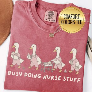 May include: A dusty rose Comfort Colors tee featuring a whimsical design of cartoon ducks dressed as nurses. The ducks are shown in various nursing roles, with the text "Busy Doing Nurse Stuff" below the illustration. A gold pendant necklace is also visible.