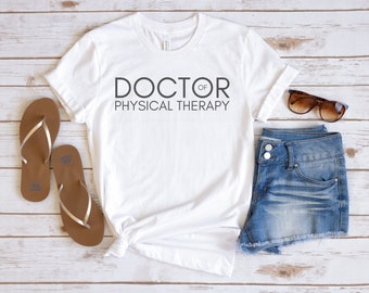 May include: White t-shirt with the text "DOCTOR OF PHYSICAL THERAPY" printed on the front. The shirt is laid flat on a wooden surface with a pair of brown flip flops and a pair of sunglasses.