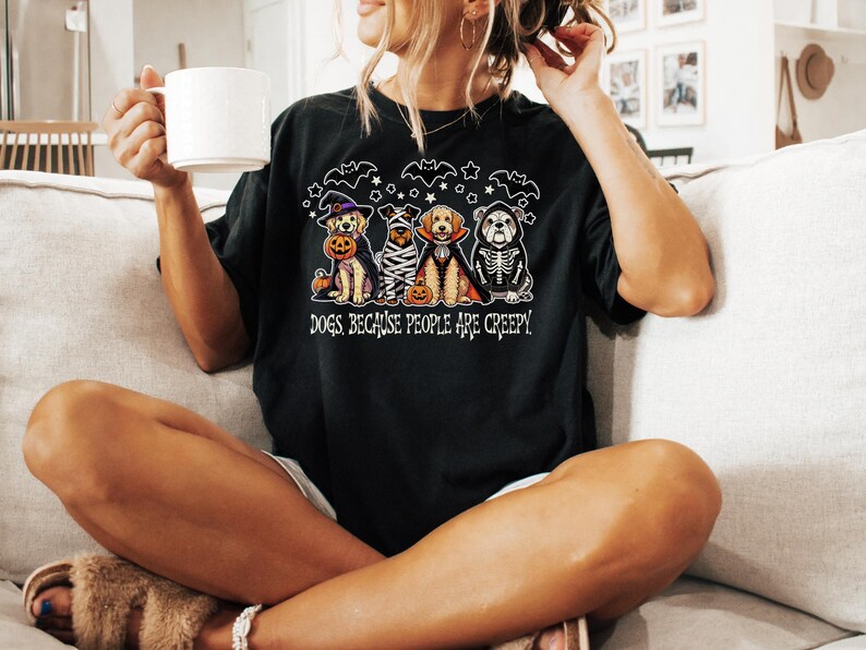 May include: Black t-shirt featuring a Halloween-themed graphic of four dogs dressed in costumes, including a witch, mummy, vampire, and skeleton. The text "DOGS BECAUSE PEOPLE ARE CREEPY" is below the dogs.