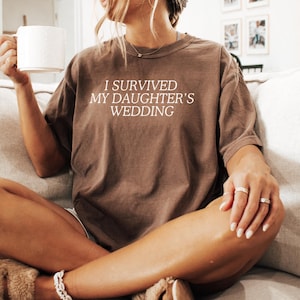 May include: A woman wearing a brown t-shirt with the text "I survived my daughter's wedding" in white. She is sitting on a sofa and holding a white mug.