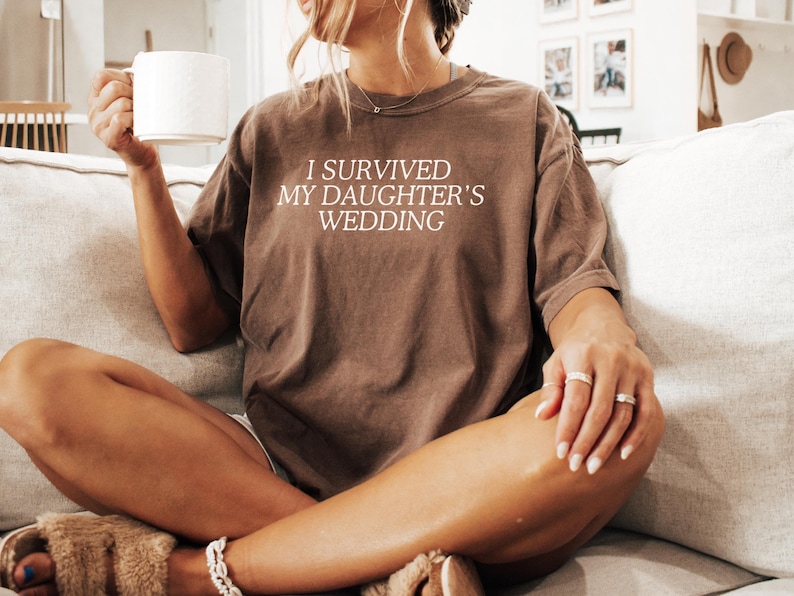May include: A woman wearing a brown t-shirt with the text "I survived my daughter's wedding" in white. She is sitting on a sofa and holding a white mug.