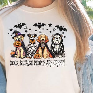 May include: Cream-colored t-shirt featuring a Halloween-themed graphic. The design includes four dogs dressed in costumes: a witch, a mummy, a vampire, and a skeleton. The text "DOGS, BECAUSE PEOPLE ARE CREEPY" is printed below the dogs.