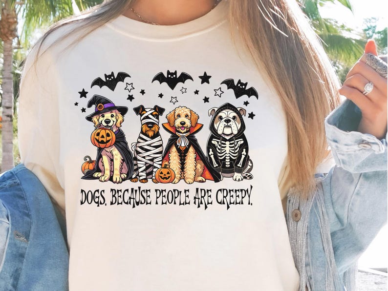 May include: Cream-colored t-shirt featuring a Halloween-themed graphic. The design includes four dogs dressed in costumes: a witch, a mummy, a vampire, and a skeleton. The text "DOGS, BECAUSE PEOPLE ARE CREEPY" is printed below the dogs.